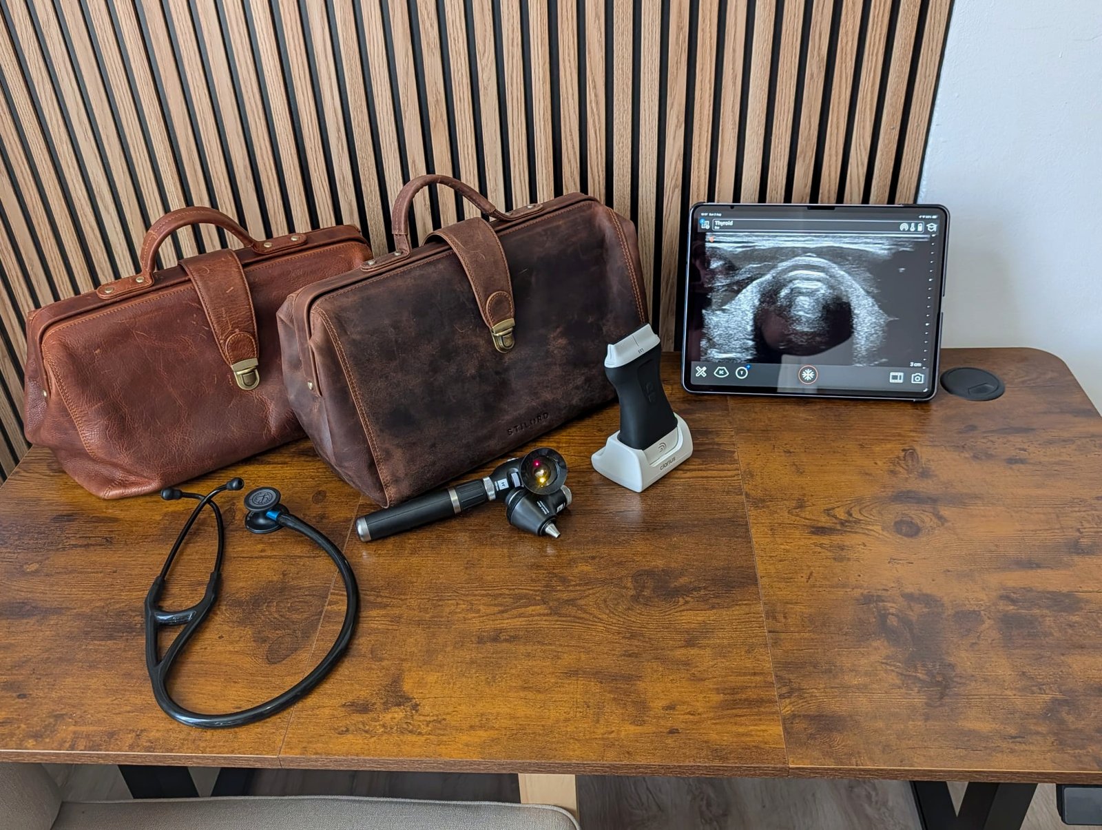 Two leather doctor bags, a stethoscope, an otoscope and ophthalmoscope set, a wireless ultrasound probe and a tablet showing a scan, on a wooden table